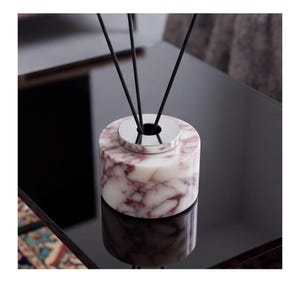 May include: A cylindrical reed diffuser base made of white marble with deep purple veining. The top features a polished silver-toned metal collar with a central opening, from which three thin black diffuser reeds extend upward.