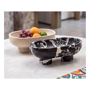 May include: Two oval-shaped footed bowls. One bowl is made of beige porous stone and contains grapes, an orange, and a lime. The second bowl is made of black and white veined marble and sits in the foreground, featuring two cylindrical feet.