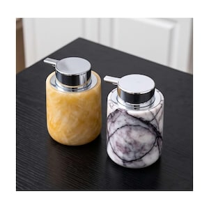 May include: Two cylindrical soap dispensers with chrome pump mechanisms. One dispenser features a translucent golden-yellow marbled pattern, while the other displays a white base with dark grey veining resembling natural stone texture.