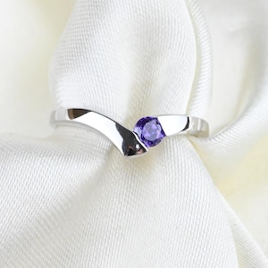 Op de afbeelding: Een zilverkleurige V-vormige ring met een enkele ronde paarse edelsteen in het midden. De ringband heeft een gladde, gepolijste afwerking en een puntig chevron-ontwerp dat naar de steen toe taps toeloopt voor een strakke uitstraling.