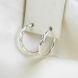 May include: A pair of small silver hoop earrings featuring a delicate diamond-cut texture along the outer surface. The earrings are attached to a rectangular white display card, showcasing a polished metallic finish and a circular hoop design.