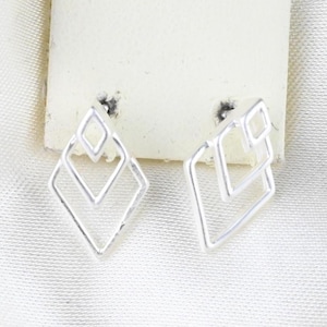 Puede incluir: Un par de pendientes de botón en tono plateado con un diseño geométrico compuesto por dos formas de diamante entrelazadas. Los pendientes están sujetos a una pequeña tarjeta de presentación rectangular blanca, con una estructura de marco abierto.