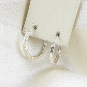Puede incluir: Un par de pendientes de aro circulares pequeños de plata con un sutil patrón diagonal grabado en la superficie metálica. Los pendientes están sujetos a una tarjeta de presentación rectangular blanca, mostrando su acabado pulido y diseño simple.