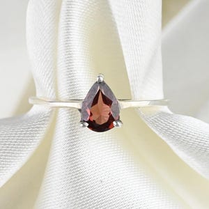 Op de afbeelding: Een zilveren ring met een enkele peervormige, dieprode granaat edelsteen gezet in een eenvoudige metalen band. De steen is gefacetteerd en wordt op zijn plaats gehouden door kleine pootjes, wat een heldere, donkerrode kleur laat zien.