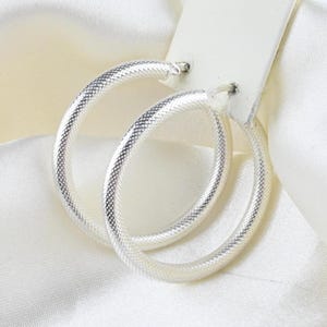 Puede incluir: Un par de pendientes de aro de plata con una superficie texturizada de patrón entrecruzado. Los pendientes circulares están sujetos a una tarjeta de presentación rectangular blanca, mostrando un acabado metálico y un diseño clásico.