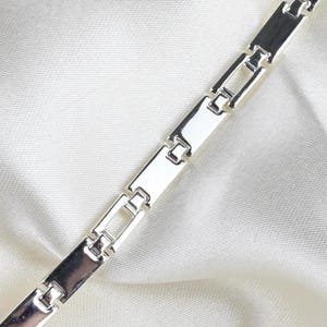 Könnte beinhalten: Ein silberfarbenes Metallarmband, das aus einer Reihe rechteckiger Glieder besteht, die durch kleinere, offene quadratische Glieder verbunden sind. Die Kette hat eine polierte, reflektierende Oberfläche und verläuft gerade.