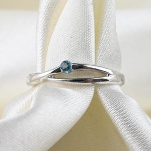 May include: A silver-toned ring featuring a split shank design that converges at the top, accented by a single small, round, light blue gemstone set centrally within the band's opening, creating a clean and polished appearance.