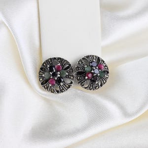 May include: A pair of round, silver-toned stud earrings. Each earring features a floral design with small, colorful gemstones in shades of red, green, blue, and black. The earrings are set against a white background.