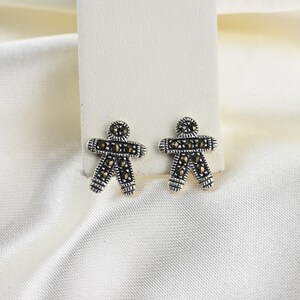 May include: A pair of sterling silver stud earrings in the shape of a person. The earrings are detailed with small, dark marcasite stones. The earrings are displayed on a white background.