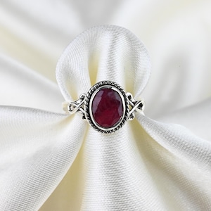 May include: A silver ring featuring a deep red oval gemstone. The gemstone is set within a textured bezel, and the band has decorative flourishes. The ring is presented on white, silky fabric, accentuating its elegant design.