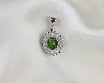 May include: A silver pendant with a central oval-cut green gemstone, surrounded by a halo of smaller, clear, rectangular stones. The pendant hangs from a small silver loop. The background is a white, textured fabric.