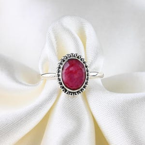 Op de afbeelding: Zilveren ring met een ovale, gefacetteerde, dieprode edelsteen. De edelsteen is omgeven door een kralenrand. De ring is geplaatst tegen een witte, getextureerde stoffen achtergrond.