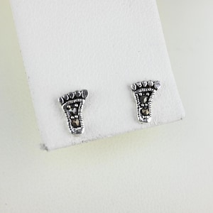 May include: A pair of silver-toned stud earrings shaped like tiny footprints. Each earring features detailed toe and arch representations, with small, dark accents. The earrings are displayed on a white surface.