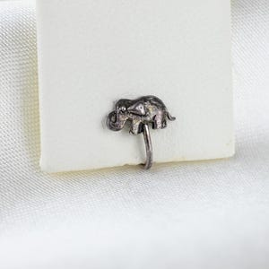 Può includere: Un piccolo anello per il naso a forma di elefante color argento. L'elefante presenta dettagli, tra cui una proboscide, orecchie e una coda. L'anello per il naso è attaccato a un perno curvo. Lo sfondo è una superficie bianca.