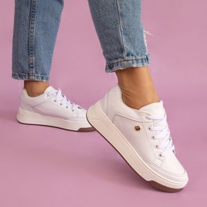Puede incluir: Zapatillas blancas de cuero con una suela gruesa blanca y un acento dorado en el lateral. Las zapatillas son usadas por una persona que lleva jeans azules.