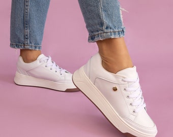 May include: White leather sneakers with a thick white sole and a gold accent on the side. The sneakers are worn by a person wearing blue jeans.