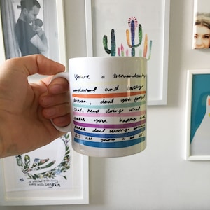 以下が含まれることがあります： 虹色のストライプ模様と「You're a tremendously wonderful person, don't you forget that. Keep doing what makes you happy and please don't worry because it's all going to be ok.」というテキストが書かれた白いセラミック製のマグカップです。