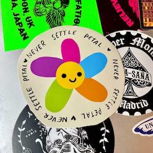 Könnte beinhalten: Ein farbenfroher Aufkleber mit einer lächelnden Blume mit rosa, blauen, orangefarbenen, lila und grünen Blütenblättern. Die Worte "NEVER SETTLE PETAL" sind um die Blume herum gedruckt. Andere Aufkleber mit Text und Grafiken sind im Hintergrund.