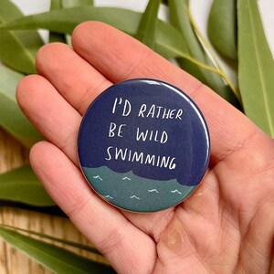 Op de afbeelding: Een ronde, marineblauwe button met de witte tekst "I'D RATHER BE WILD SWIMMING". De onderkant van de button heeft een wit golfontwerp op een turquoise achtergrond. De button wordt in een hand gehouden.