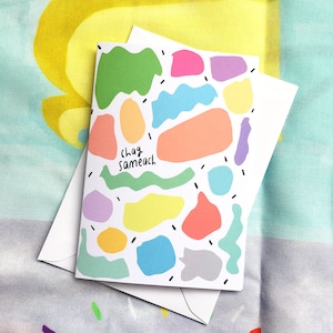 Könnte beinhalten: Eine weiße Karte mit einem bunten abstrakten Design und dem Text "clag sameach" in Grün und Schwarz. Die Karte ist von einem weißen Umschlag umgeben.