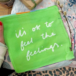 以下が含まれることがあります： 白い筆記体で「it's ok to feel the feelings」と書かれた、正方形の鮮やかな緑色の生地。生地は人によって保持されています。背景には花柄とヒョウ柄が含まれています。