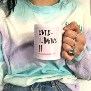 Peut inclure: Une tasse en céramique blanche avec une bande rose et du texte noir qui dit "OVER-THINKING IT".