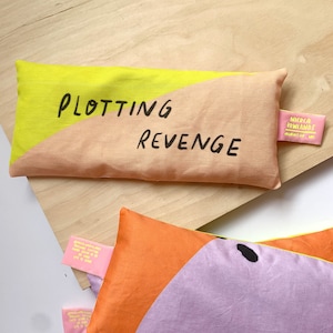 May include: A yellow and peach-colored eye pillow with the text "Plotting Revenge" in black. The pillow has a small pink label with the text "Nicola Rowlands" and a website address.