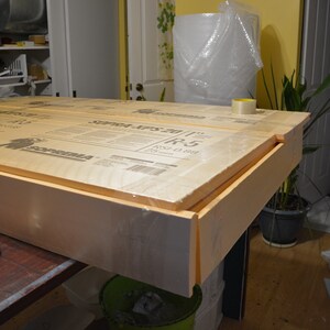 May include: A rectangular wooden frame with a foam core, wrapped in clear plastic. The frame is on a dark surface, with a measuring tape visible. The product appears to be construction material.
