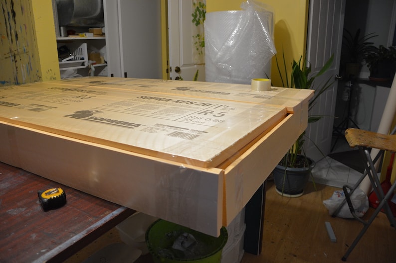 May include: A rectangular wooden frame with a foam core, wrapped in clear plastic. The frame is on a dark surface, with a measuring tape visible. The product appears to be construction material.
