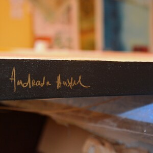May include: Detail of a canvas artwork with a black border. The artist's signature, "Andrada Hustul," is inscribed in gold script on the side. The painting's colours are visible in the background.