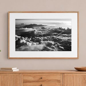 Puede incluir: Una fotografía de paisaje en blanco y negro de una densa formación de nubes vista desde arriba, con rayos de luz y sombras dramáticas, exhibida en un marco de madera rectangular con un borde de paspartú blanco ancho.