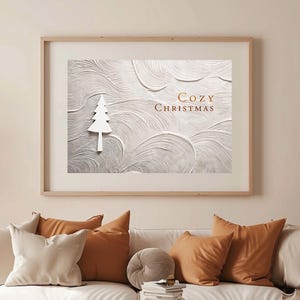 Puede incluir: Una impresión de arte mural horizontal enmarcada que presenta un fondo texturizado de color blanco roto con patrones de remolinos, una silueta blanca de un pino y el texto "COZY CHRISTMAS" en una fuente de cobre metálico sobre un sofá blanco.