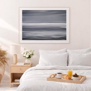 Puede incluir: Una impresión artística abstracta enmarcada que representa una escena oceánica en escala de grises. La obra de arte cuelga sobre una cama con ropa de cama blanca. Una mesita de noche de madera con una lámpara y flores está al lado de la cama.