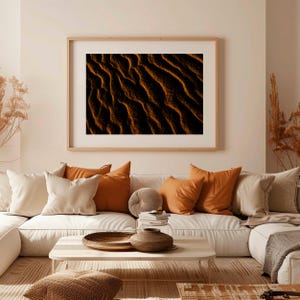 Puede incluir: Una impresión artística enmarcada que muestra un primer plano de dunas de arena en tonos cálidos de naranja y negro. La obra de arte cuelga sobre un sofá blanco adornado con cojines naranjas y beige, creando un ambiente acogedor.