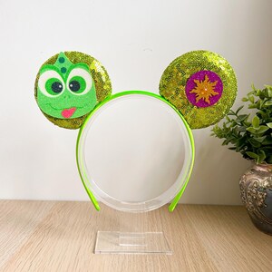 Peut inclure: Serre-tête vert citron avec deux oreilles rondes à paillettes. Une oreille représente Pascal, un personnage vert, l'autre un soleil violet et doré. Le serre-tête est présenté sur un support transparent.