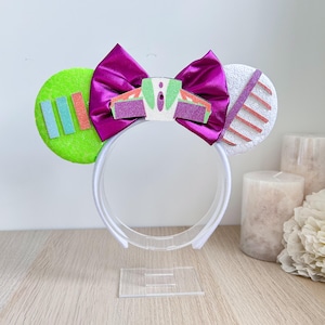 May include: A white headband with attached mouse ears. One ear is lime green with blue and orange rectangles. The other ear is white with pink and orange stripes. A large, magenta bow sits between the ears. The headband is displayed on a clear stand.