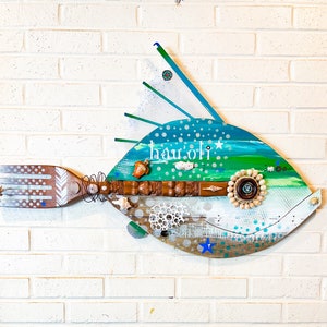 May include: A blue, green and white fish-shaped wall art piece with the word "hauloli" written on it. The fish is made of wood and has a variety of textures and embellishments, including shells, beads and a star.