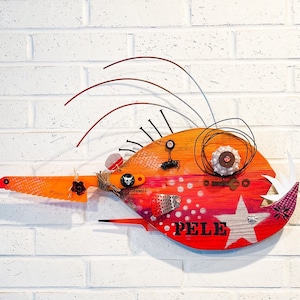 Puede incluir: Escultura de pez de madera hecha a mano, con una combinación de colores naranja, rojo y blanco. La obra de arte incluye clavos, alambre y una estrella, con el nombre "PELE" impreso. El pez está montado en una pared de ladrillos blancos.