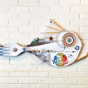 Könnte beinhalten: Eine weiße Holzfischskulptur mit einer blau-weißen Gabel als Schwanz. Der Fisch ist mit verschiedenen Verzierungen versehen, darunter Muscheln, ein Stern, ein gewebter Kreis und ein Regenbogenaufkleber mit der Aufschrift "Hawaii".