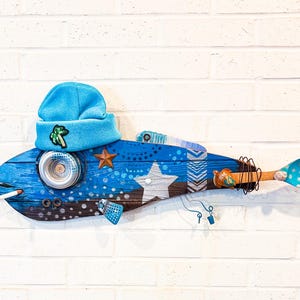 May include: A quirky, handmade wooden fish sculpture, painted blue with white details and star motifs. It has a blue beanie with a green badge, a metal can eye, and a wooden spoon tail. The fish is mounted on a white brick wall.