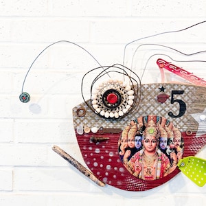 May include: Mixed media artwork on a white brick wall. The artwork features a red and tan wooden base with a collage of elements, including a bottle cap, shells, and a religious image. The number "5" is prominently displayed.