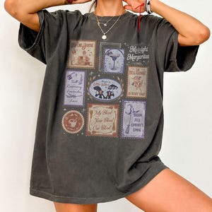May include: A charcoal gray short-sleeve t-shirt featuring a grid of rectangular graphic prints. The designs include illustrations of a house, a cocktail, a witch hat, and text such as Midnight Margaritas, Salt Rosemary Lavender, and My Blood Your Blood.