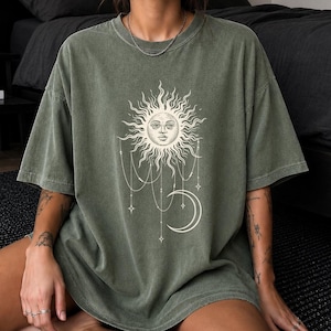 May include: An olive green crew neck t-shirt featuring a large, cream-colored graphic print on the chest. The design depicts a stylized sun with a human face, surrounded by radiating rays, with dangling chains, small stars, and a crescent moon below.