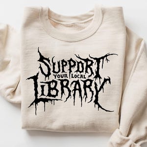 May include: A cream-colored crewneck sweatshirt featuring black graphic text on the front that reads SUPPORT YOUR LOCAL LIBRARY in a stylized, jagged, and dripping font. The garment has long sleeves and a ribbed collar, cuffs, and hem.