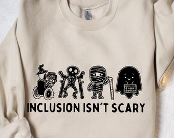 May include: Beige sweatshirt with a black graphic of Halloween-themed characters, including a witch in a wheelchair, a skeleton, a mummy, and a ghost. The text "INCLUSION ISN'T SCARY" is printed below the characters. The sweatshirt is folded on a white surface.