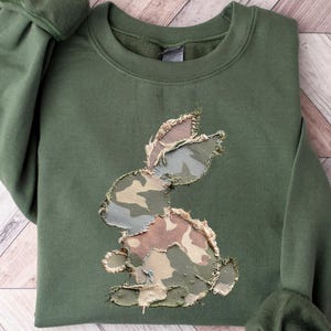 May include: An olive green sweatshirt featuring a camouflage bunny applique. The bunny design is made of various patches of camouflage fabric. The sweatshirt has a crew neck and long sleeves with ribbed cuffs. The garment is laid flat on a light wood-grain surface.