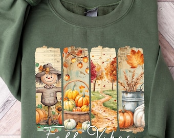 May include: An olive green sweatshirt featuring a fall-themed graphic. The graphic includes a scarecrow, pumpkins, and autumn foliage. The words "Fall Vibes" are written in a cursive font across the bottom of the graphic.