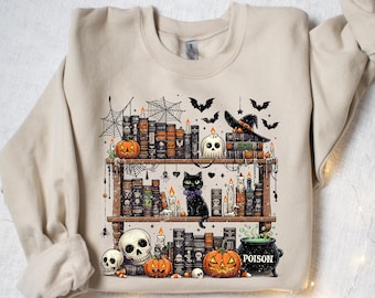 May include: Beige sweatshirt featuring a Halloween-themed graphic. The design includes a bookshelf with books, a black cat, pumpkins, skulls, candles, bats, and a witch's hat. A cauldron with the word "POISON" is also visible.