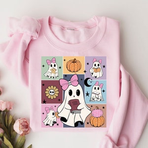 May include: A light pink sweatshirt featuring a colorful graphic of cartoon ghosts with pink bows, pumpkins, and a sunflower. The ghosts are reading books. The design is set against a grid of various colors.