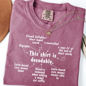 May include: A mauve short-sleeved t-shirt with white text that reads: This shirt is decodable, surrounded by arrows pointing to linguistic notes including Closed Syllable, Digraphs, r-controlled, and Latin-based roots. The shirt has a crew neckline.
